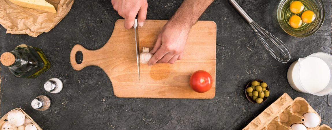 A cutting board on which a mushroom is cut. There is a marble background with olive oil, salt, pepper, eggs, olives and milk