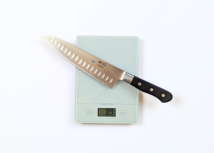 The Mac Professional 8 Inch Hollow Edge Chef Knife, diagonal on a white background while weighted on a white digital scales at 6.49 ounces